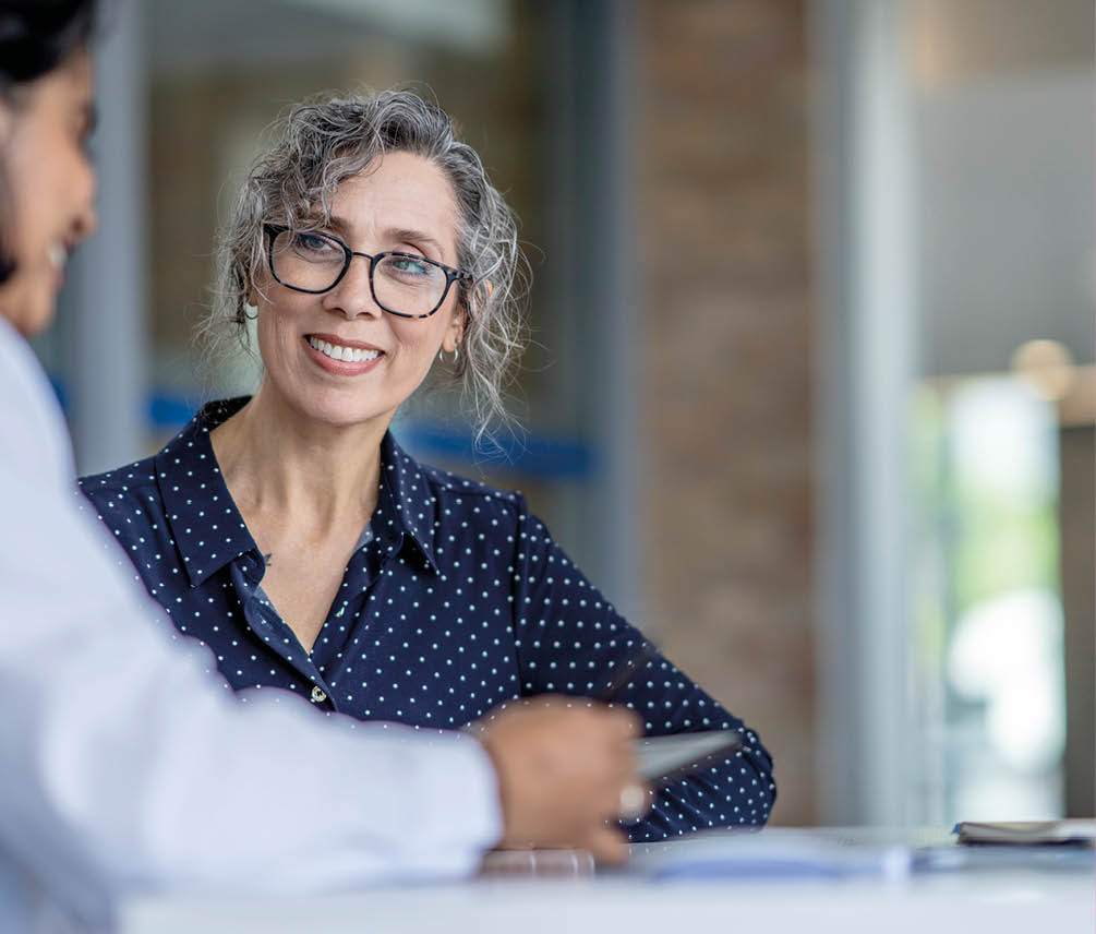 A middle aged woman sits with her female doctor of Middle Eastern decent as they review her medical concerns. She is dressed semi-casually and sharing with the doctor her recent discomfort.  The doctor is wearing a white lab coat and taking notes on her tablet as the two talk.